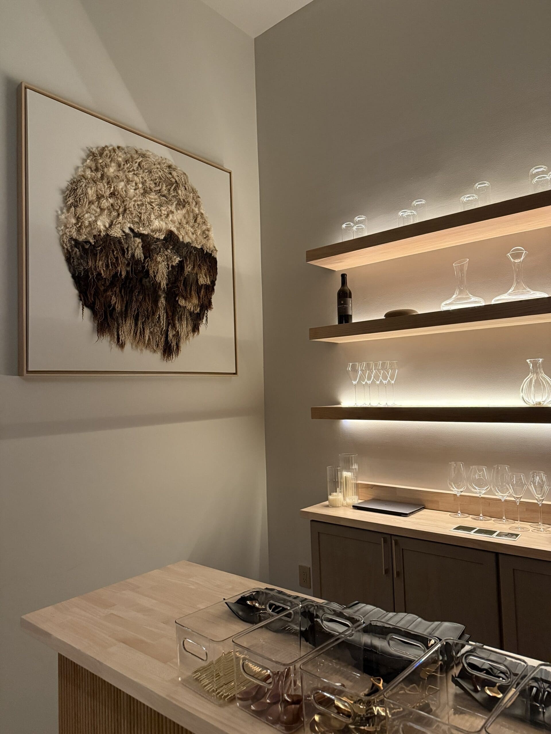 a Modern Home Bar with Wooden Shelves Holding Glassware Decanters and a Bottle Softly Lit from Below on the Wall is a Large Textured Artwork Made of Layered Feathers in Natural Tonesperfect for a Burnt Hill Farm Inspired Retreat