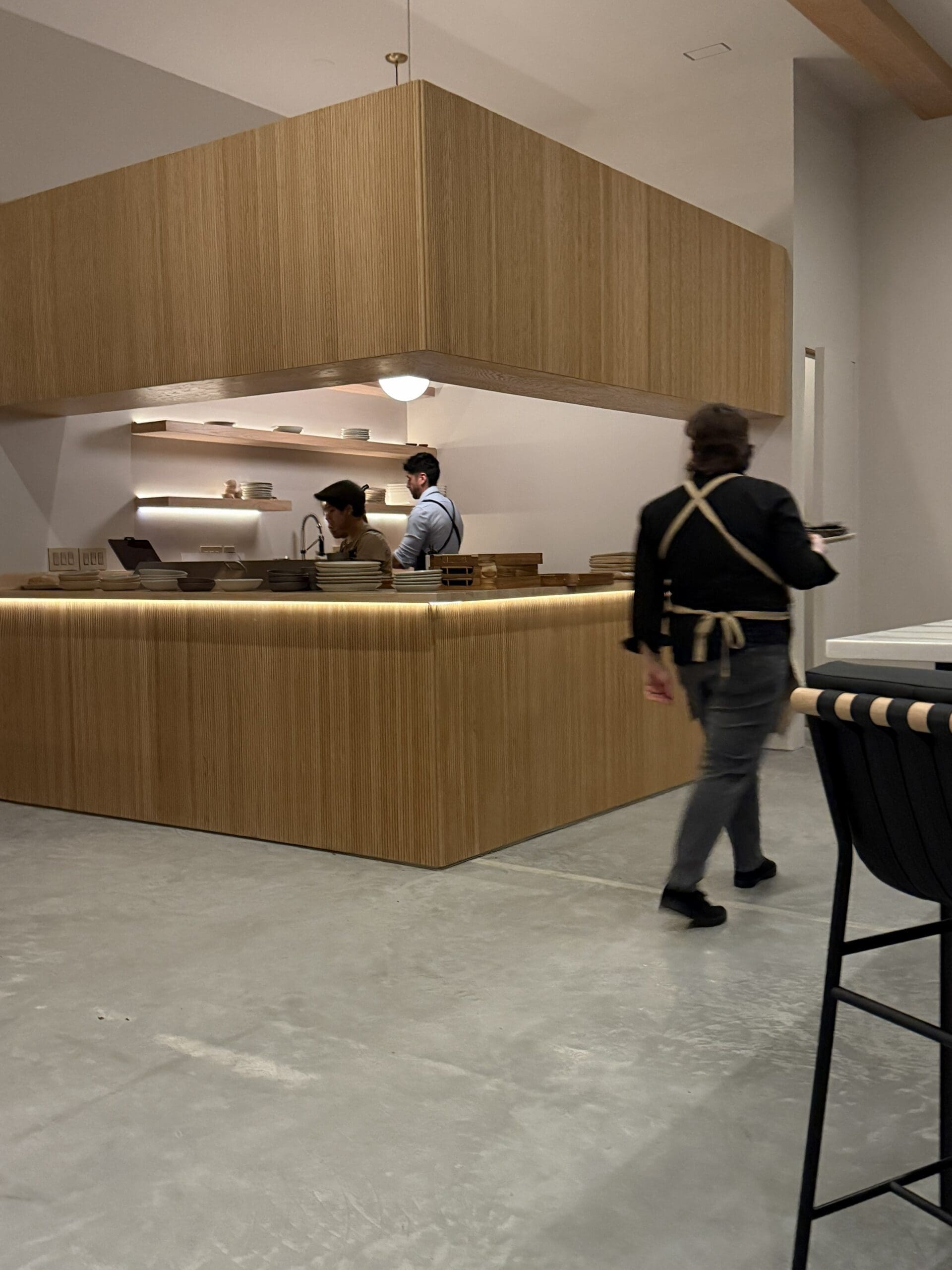 a Modern Minimalist Café with Light Wood Panels and Burnt Hill Farm Coffee Two Staff Behind the Counter Work on Orders One Masked an Apron Clad Person Walks by with a Notepad the Floor is Concrete Barstools Line the Counter in the Foreground