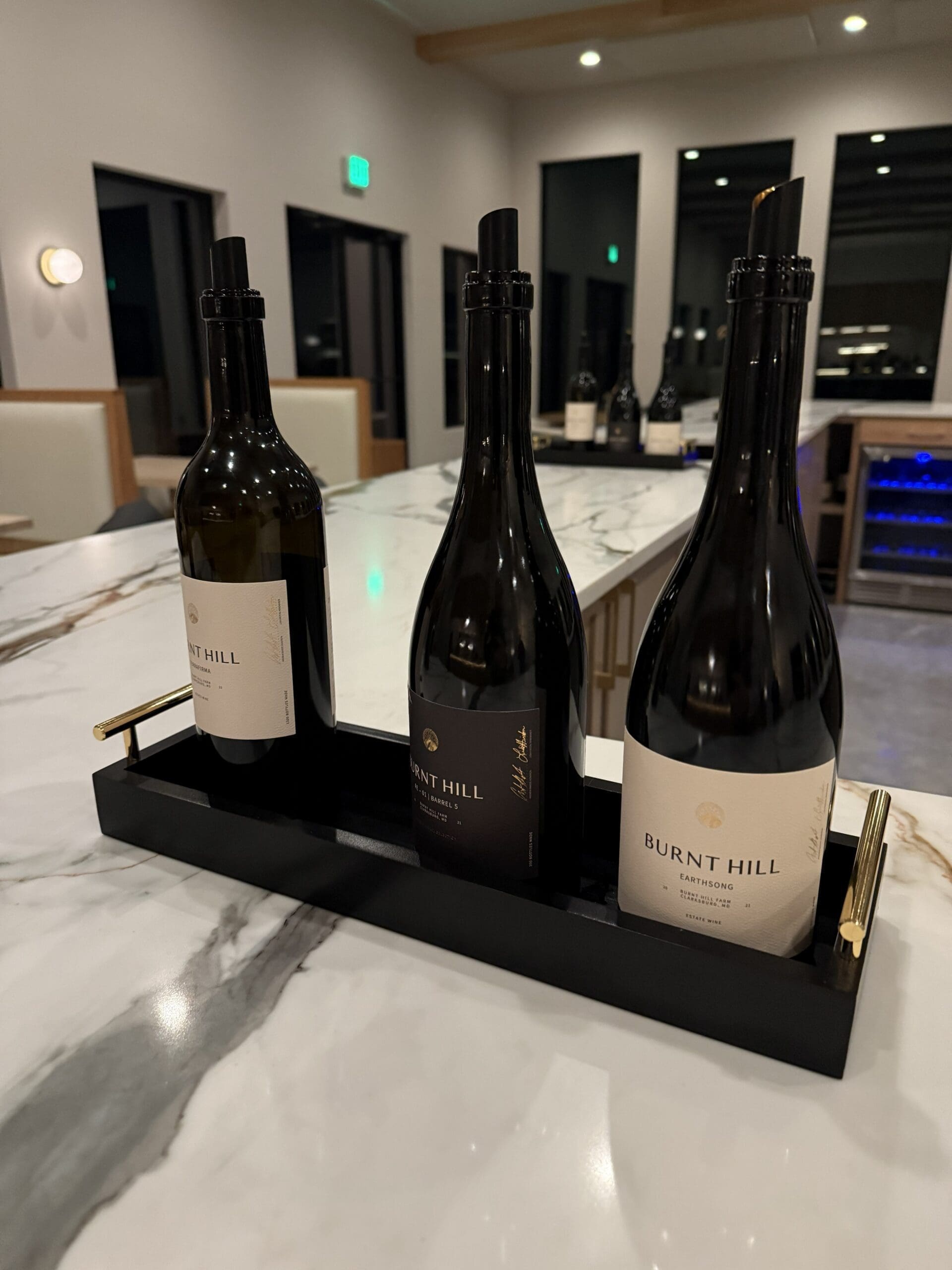 Three Burnt Hill Wine Bottles Are Displayed Upright in a Sleek Black and Gold Tray on a Marble Countertop the Modern Burnt Hill Farm Tasting Room Features Beige Seating Large Windows and More Wine Bottles on Another Counter