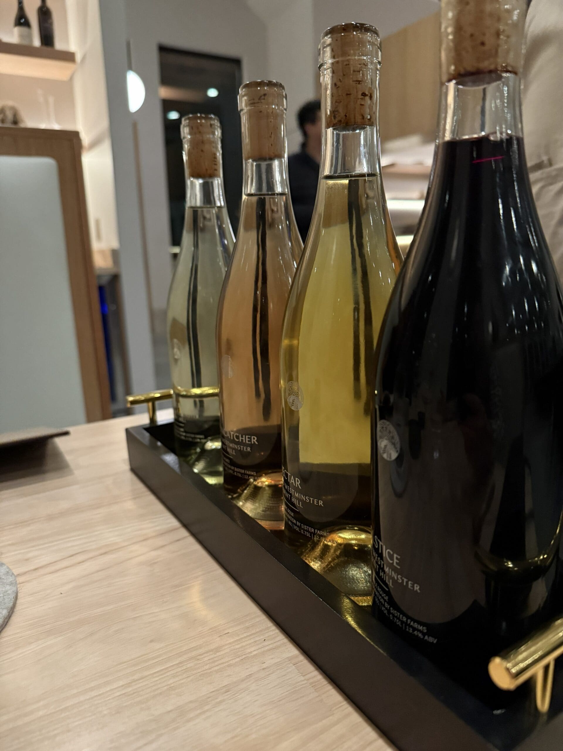 Four Wine Bottles with Cork Stoppers from Burnt Hill Farm Are Displayed in a Black Tray with Gold Handles on a Light Wooden Table the Bottles Hold Liquids in Shades from Clear to Amber to Deep Red with a Blurred Indoor Setting and People in the Background