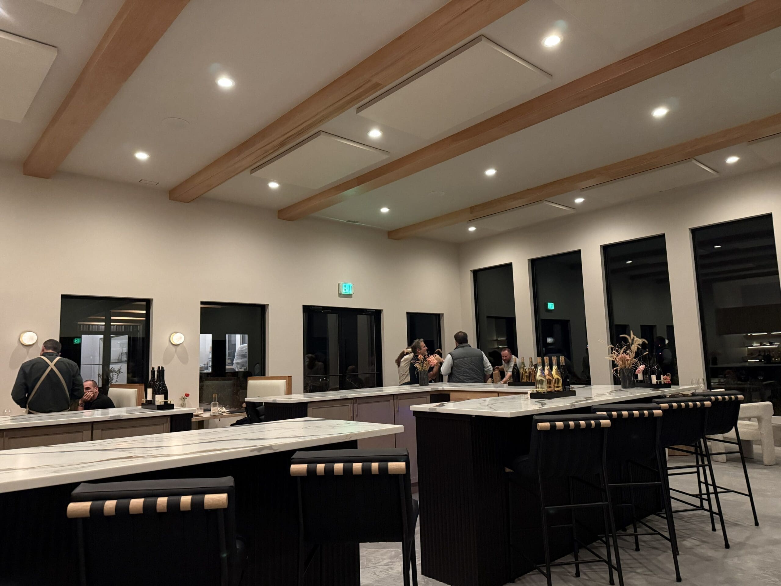 a Modern Well Lit Bar with High Black Chairs Marble Countertops and Exposed Wooden Beams Guests Sip Burnt Hill Farm Wines and Converse by the Bar Bottles Glasses Minimalist Decor and Large Windows Complete the Inviting Scene