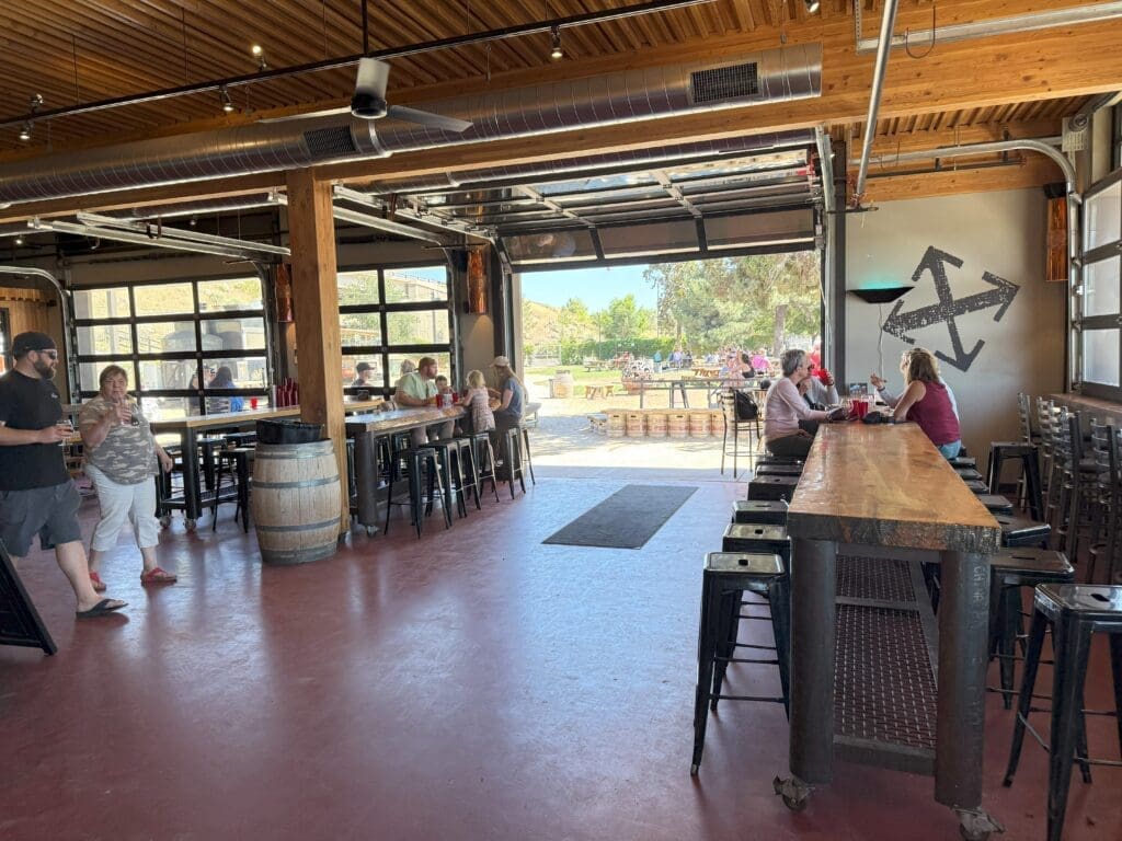 a Bright Open Taproom at Crux Fermentation Project Features Wooden Beams High Ceilings and Garage Doors Opening to a Sunny Patio Guests Relax at Tall Tables and the Long Bar Under a Bold Black Arrow Mural with Outdoor Seating Visible Through the Doors