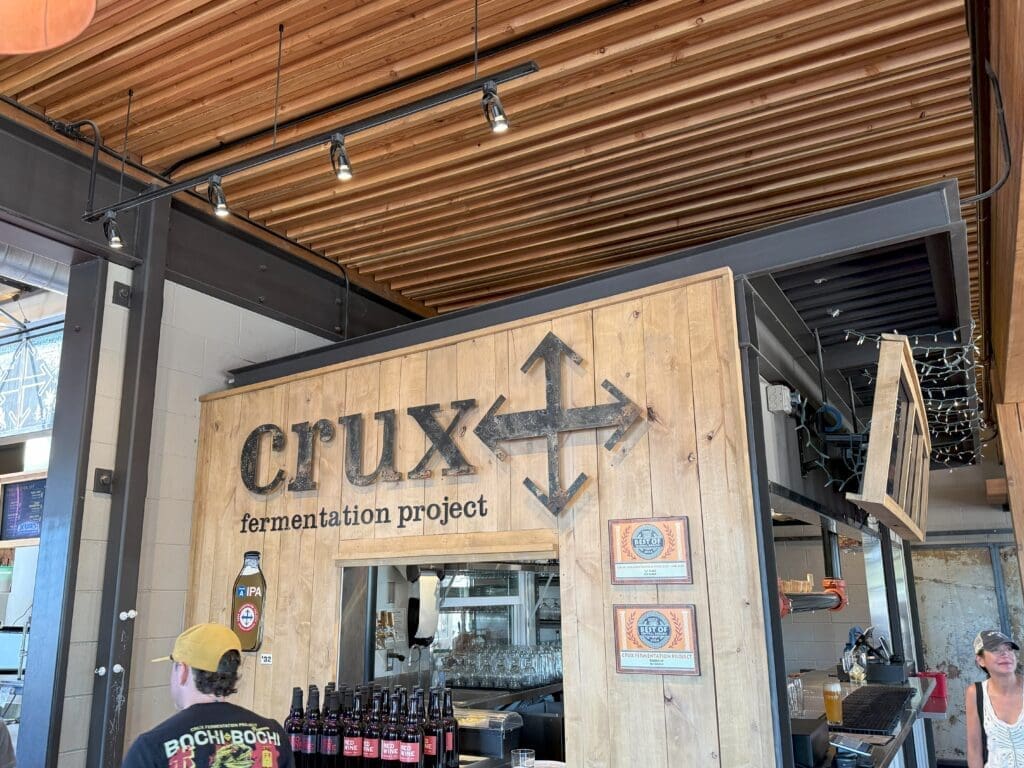 a Small Wooden Booth Labeled Crux Fermentation Project Features a Bold Black Arrow Logo Beer Bottles on Display and Several Awards a Staff Member in a Cap Stands by As Bar Glasses Hang Inside with Exposed Beams and Ceiling Overhead