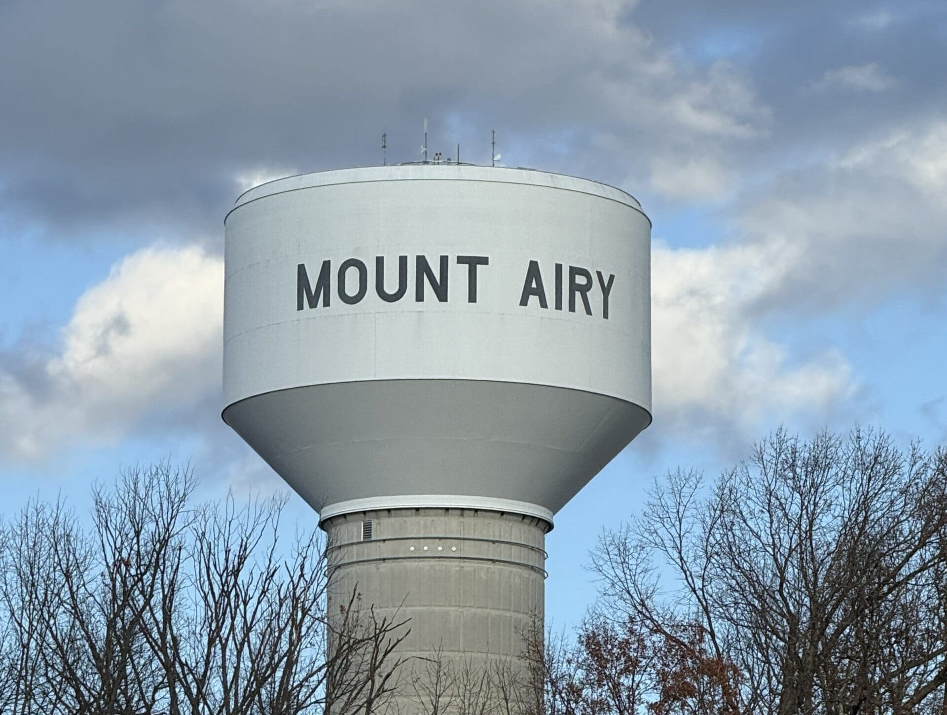 A tall, light gray water tower with MOUNT AIRY in bold black letters rises above leafless trees and a partly cloudy sky—a familiar sight for those exploring things to do in Frederick, MD. The tower’s cylindrical base supports its rounded top.