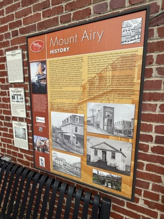 an Informational Sign Titled Mount Airy History is Mounted on a Red Brick Wall Above a Black Metal Bench the Sign Features Black and White Photos and Text About Mt Airy Mds Past Including Its Old Buildings and Town Events