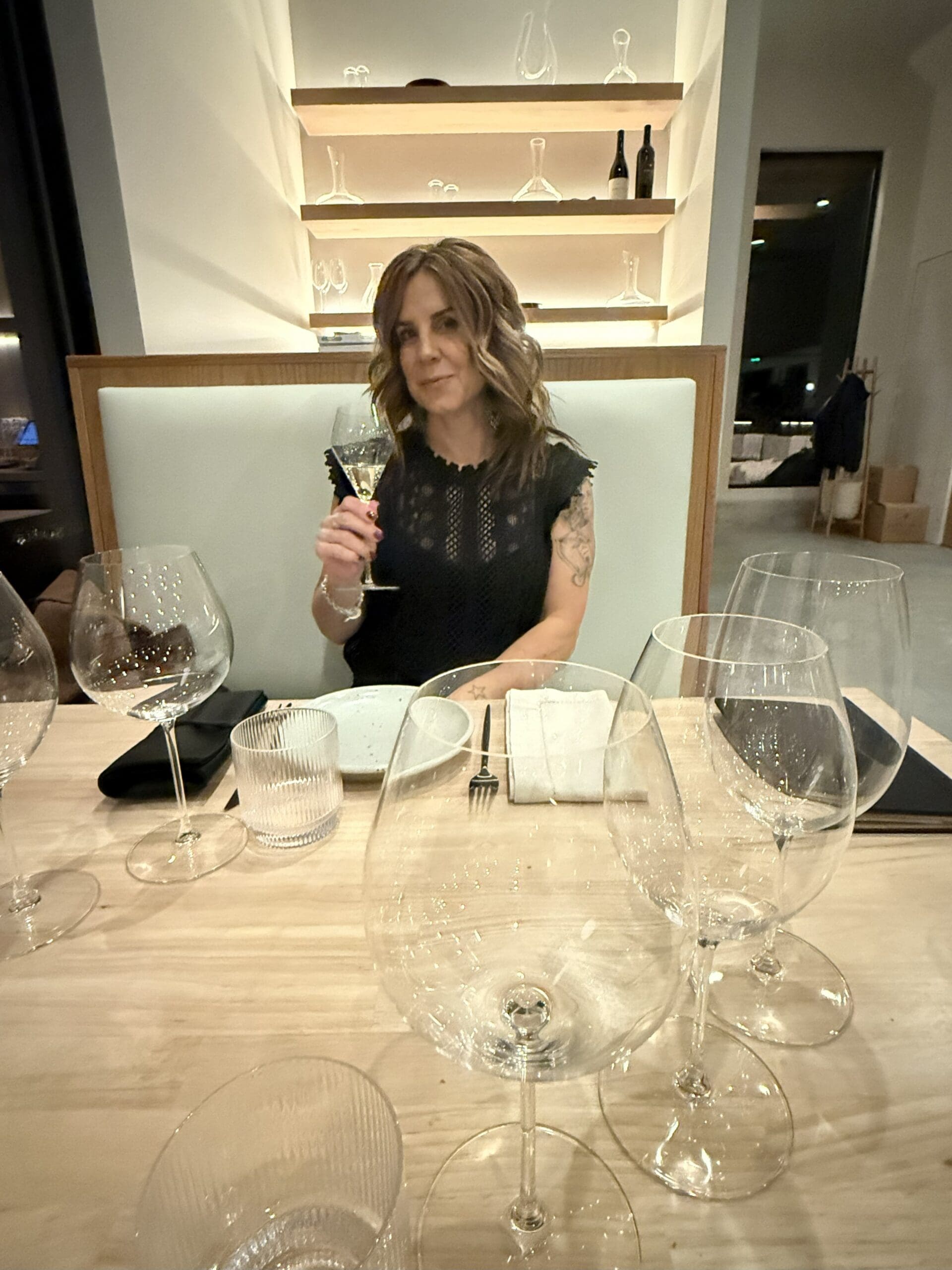a Woman with Wavy Brown Hair in a Black Dress Sits at a Restaurant Table Holding a Wine Glass from Burnt Hill Farm She is Smiling Slightly the Table Has Several Empty Wine Glasses Plates and Cutlery Shelves with Glassware Are on the Wall Behind Her