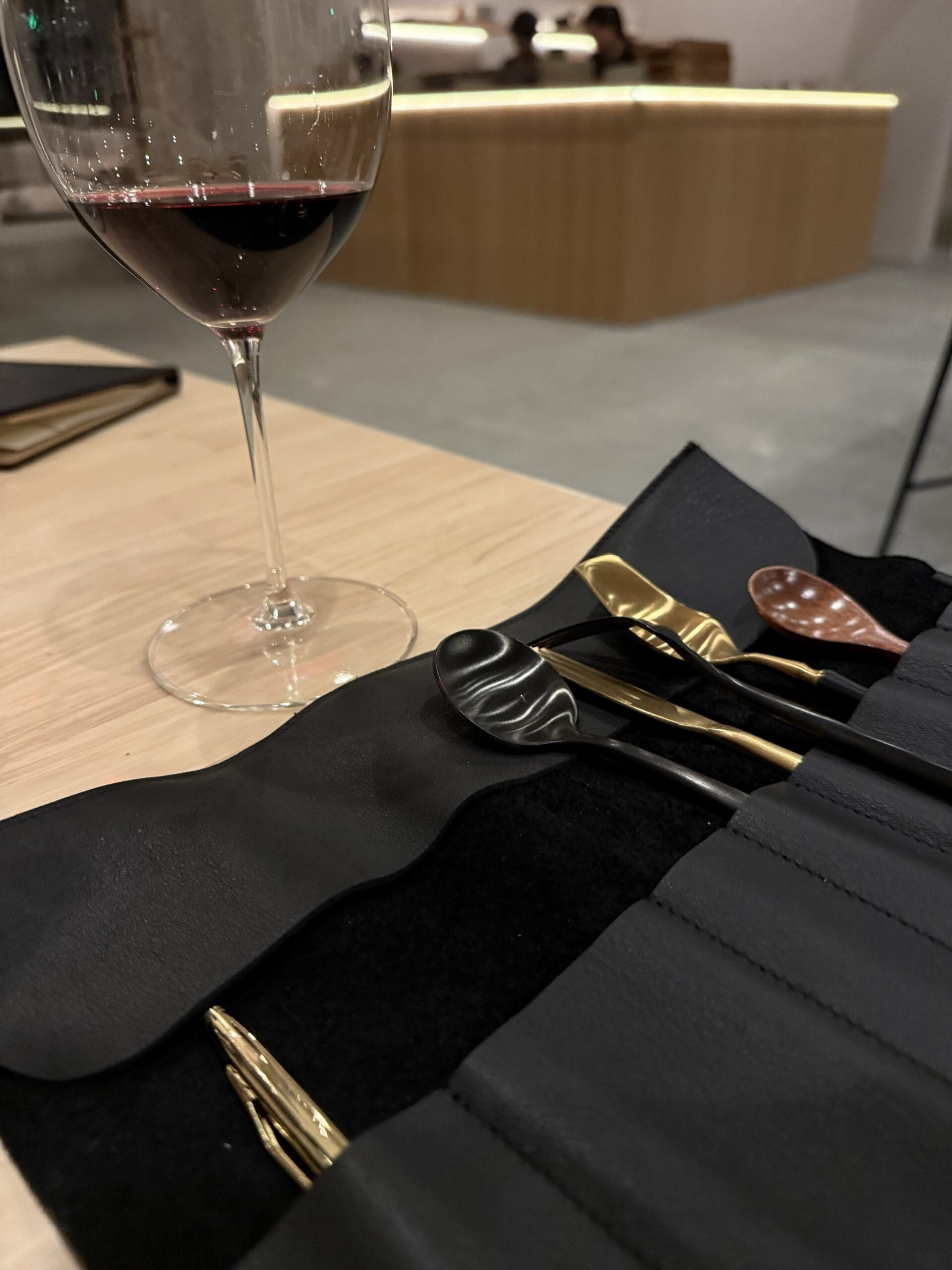 a Glass of Red Wine Sits on a Light Wood Table Next to a Black Case Holding Elegant Metal Utensils with Colored Handles Set in a Modern Minimalist Restaurant the Ambiance Hints at the Refined Flavors of Burnt Hill Farm