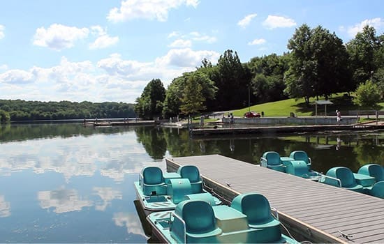 a Calm Lake with Pedal Boats Docked by a Wooden Pier in the Foreground Near Mt Airy Md Trees and Grassy Hills Surround the Water with Blue Skies and Clouds Reflected on Its Surface People Stroll Along the Distant Shoreline