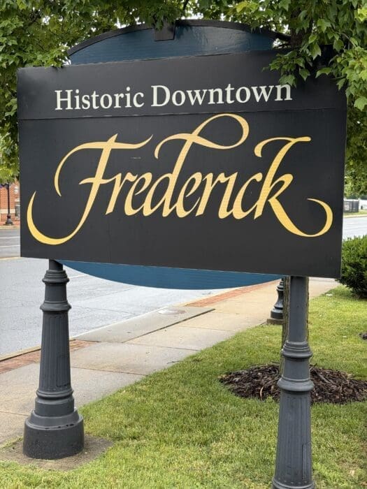 A large black sign with ornate yellow script reads Historic Downtown Frederick. Supported by two decorative posts, the sign is surrounded by grass, mulch, and overhanging green branches—highlighting EpicFrederick's dedication to community partnership.