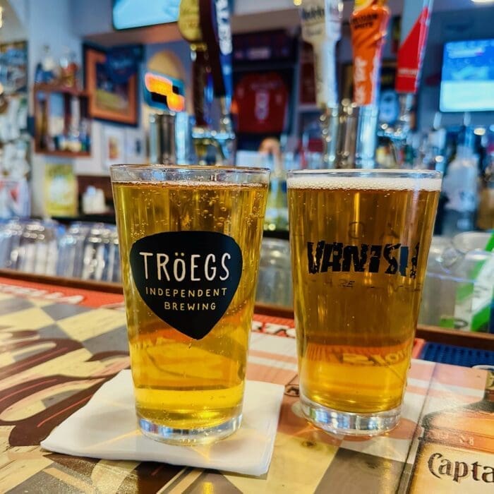Two pint glasses of golden beer sit on a bar counter—one with a Tröegs Independent Brewing logo, the other Vanish. Both rest on white napkins. Behind them are beer taps and blurred lights, perfect for enjoying after pizza in Frederick MD.