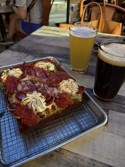 A metal tray holds thick, square slices of Detroit-style pizza in Fredrick MD, topped with pepperoni, cheese, and herbs. Next to it are two pints of beer—one light and one dark—on a wooden table with people seated in the background.