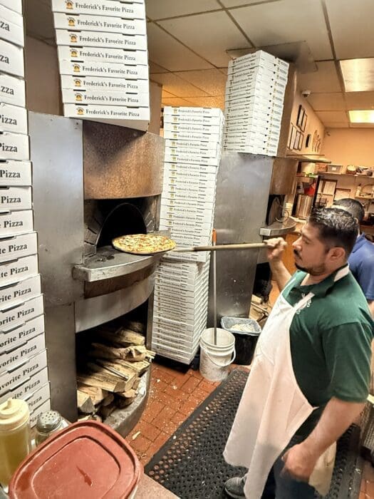 A man in a green shirt and white apron uses a long pizza peel to remove pizza in Frederick MD from a wood-fired oven. Stacks of “Frederick’s Favorite Pizza” boxes surround him, with firewood stored below the oven inside a busy pizzeria kitchen.
