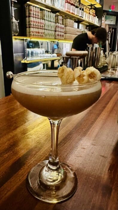 A creamy cocktail garnished with popped popcorn sits in a coupe glass on a wooden bar counter at Cucina Massi Frederick MD. Warm lighting and shelves lined with bottles create a cozy atmosphere as a bartender works in the background.