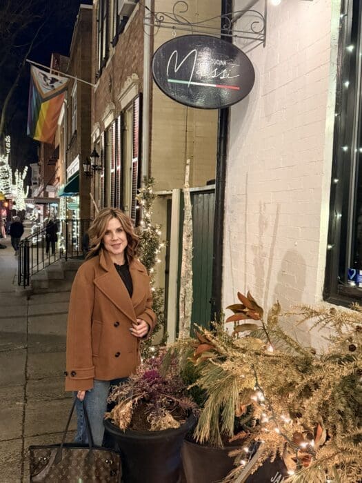A woman in a brown coat stands smiling on a lit city sidewalk at night beside winter greenery. Behind her, a Cucina Massi Frederick MD sign glows as festive lights and rainbow flags decorate the welcoming street.
