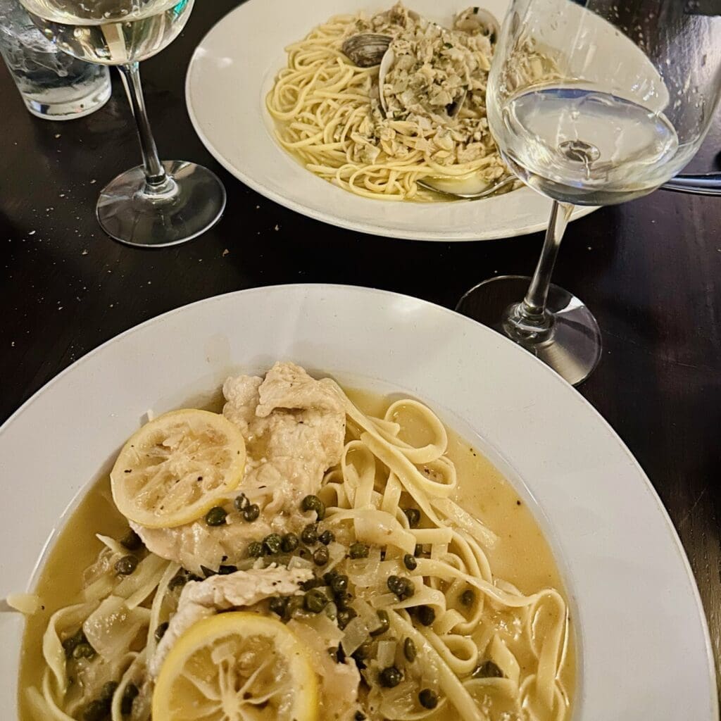 Two plates of pasta on a dark table at Cucina Massi Frederick MD: front dish features linguine with lemon, chicken, capers, and sauce; back dish has linguine with clams. Two glasses of white wine and a glass of water are also visible.