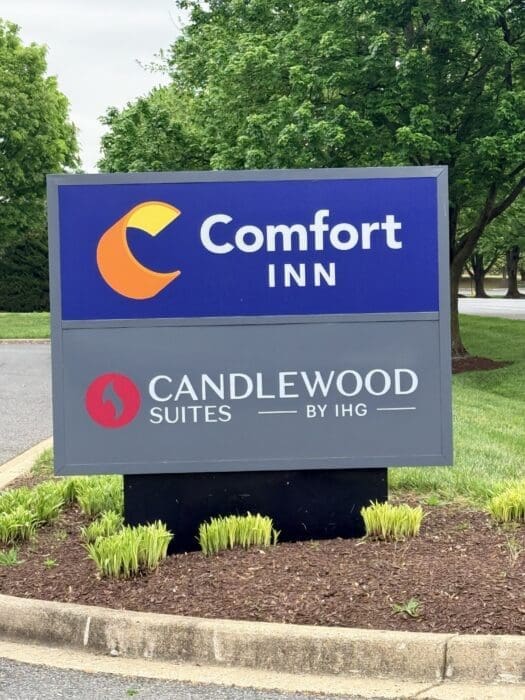 Blue Comfort Inn sign with orange crescent logo, advertising Candlwood Suites by IHG, set among green trees and landscaped ground. Some of the best cheap hotels in Frederick, MD.