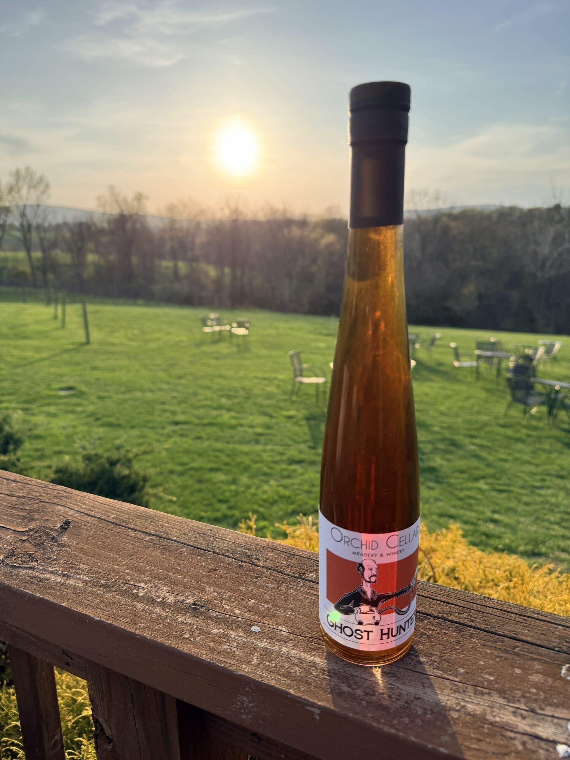 a Tall Slender Bottle of Orchid Cellar Wine Stands on a Wooden Railing in Front of a Green Lawn and Scattered Patio Furniture with Trees and Hills in the Distance Under a Setting Sun at Orchid Cellar Middletown Md