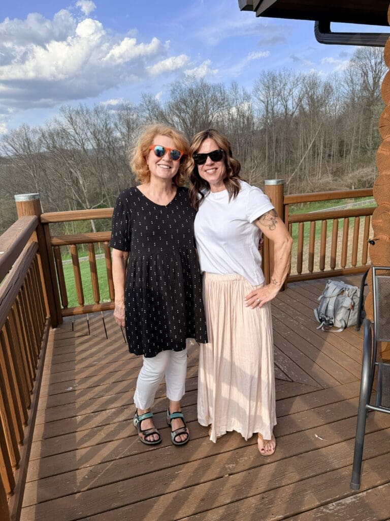 Two Women Stand Smiling on a Wooden Deck at Orchid Cellar Middletown Md Both Wear Sunglasses and Sandals with Trees and a Grassy Area in the Background Under a Partly Cloudy Sky