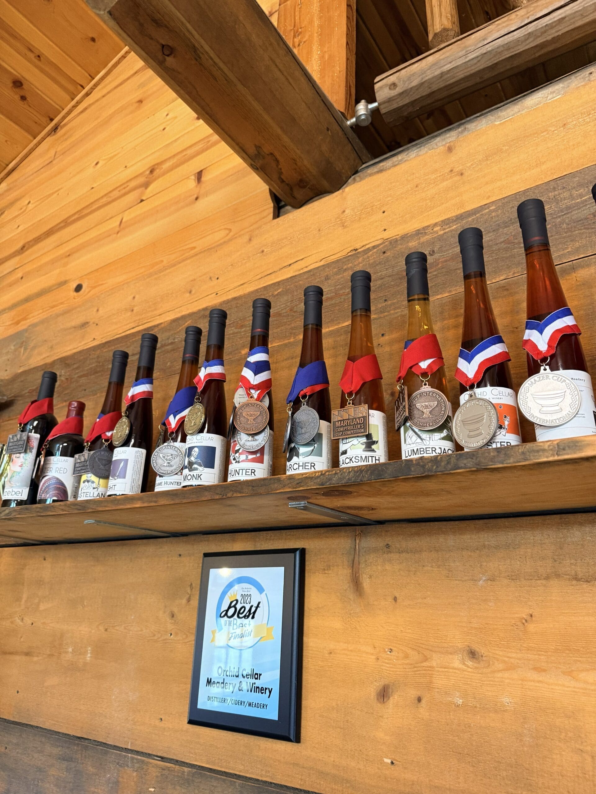 a Row of Tall Wine Bottles with Medals Hangs on a Wooden Shelf Against a Wooden Wall Below a Framed 2023 Best of the Best Award Plaque for Orchid Cellar Middletown Md is Displayed Honoring Their Winery in Ottawa County