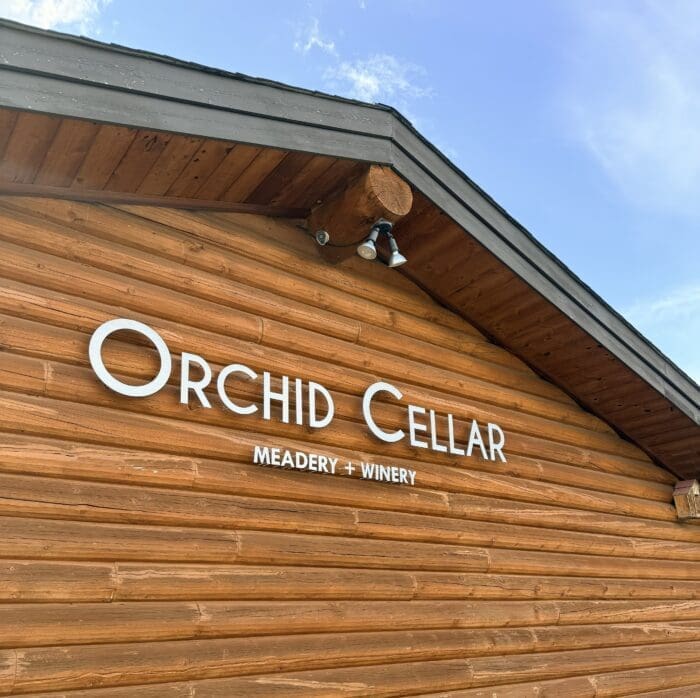 The image shows a wooden building with a sloped roof against a blue sky in Frederick, Maryland. On the wood siding, large white letters read Orchid Cellar and below in smaller text, Meadery + Winery. Two outdoor lights are mounted above the sign.