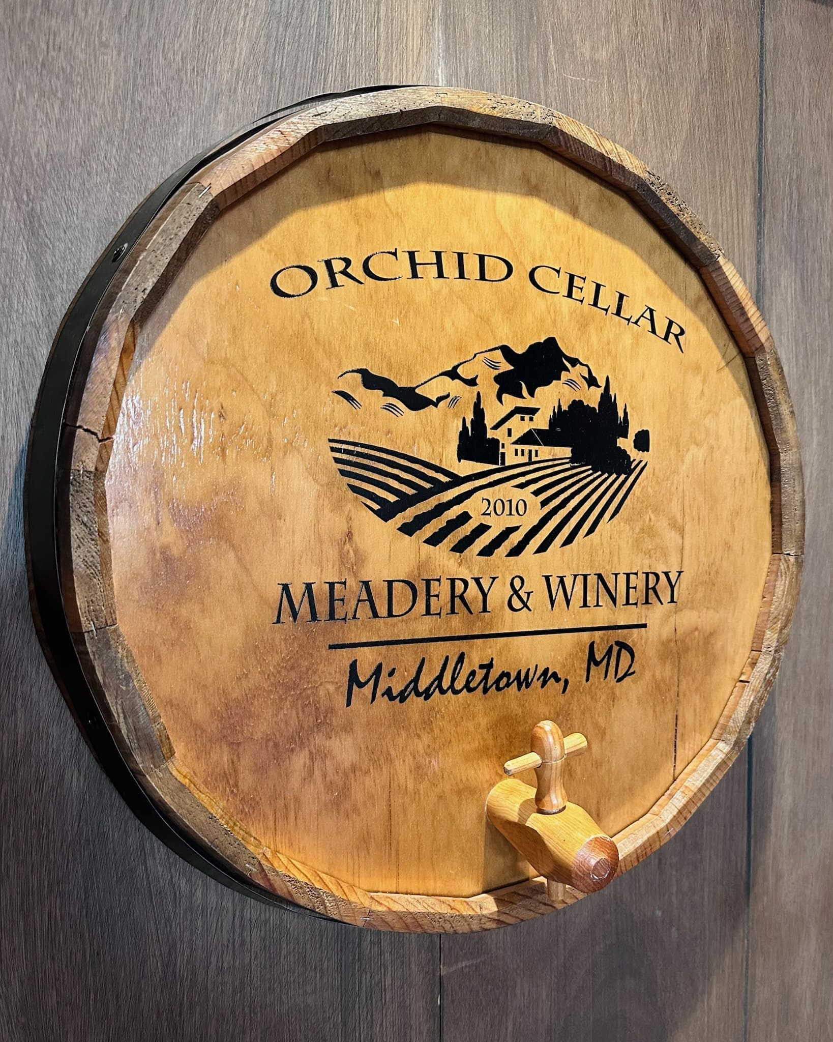a Round Wooden Barrel Lid with a Spout Mounted on a Wood Wall Displays orchid Cellar Middletown Md 2010 and a Logo of Hills a House Vineyards and Birdsshowcasing the Unique Spirit of Orchid Cellar