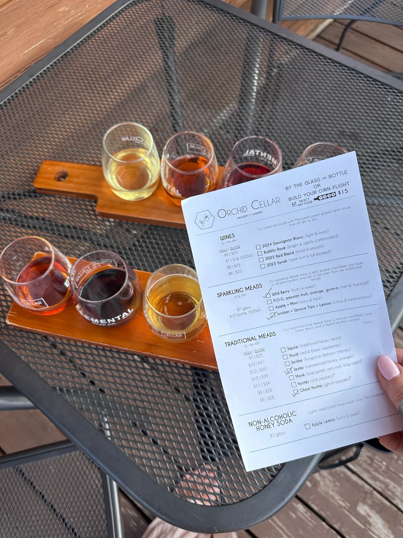 a Hand Holds a Tasting Menu Above Two Wooden Trays Each with Four Small Glasses of Dark Amber and Golden Meads from Orchid Cellar Middletown Md the Trays Rest on a Black Metal Mesh Table Outdoors Sunlight Casting Shadows Across the Surface