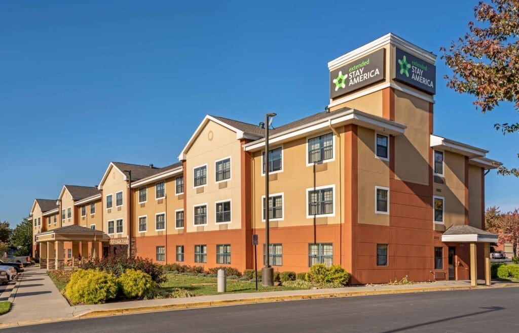 Exterior of an Extended Stay America hotel with tan and orange walls, multiple windows, and a clear blue sky above.