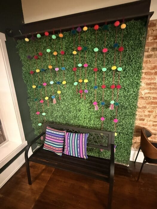Black Bench Against a Faux Greenery Wall Decorated with Vertical Strings of Multicolored Pom Poms and Two Colorful Striped Pillows
