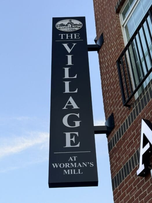 Vertical Black Sign Reading the Village at Wormans Mill with a Small Oval Logo of a Man in a Landscape Mounted on a Brick Wall in Frederick Md