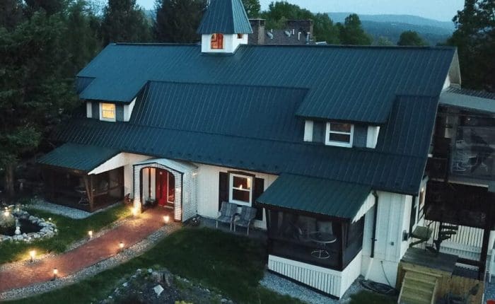 A large, dark-roofed house with dormer windows and a central peaked tower, resembling the charm of historic hotels in Frederick MD. It features screened porches, garden beds, a red-brick walkway, and green trees under a cloudy sky.
