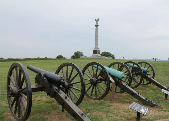 Row of old cannons on a grassy parade ground with a tall monument featuring an eagle statue in the distance