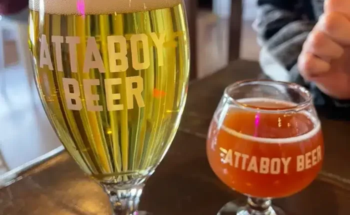 Two glasses of beer on a wooden table feature “ATTABOY BEER” branding, showcasing the vibrant scene of local breweries Frederick offers. A pale golden beer and an amber brew highlight the variety at one of the top breweries in Frederick MD.