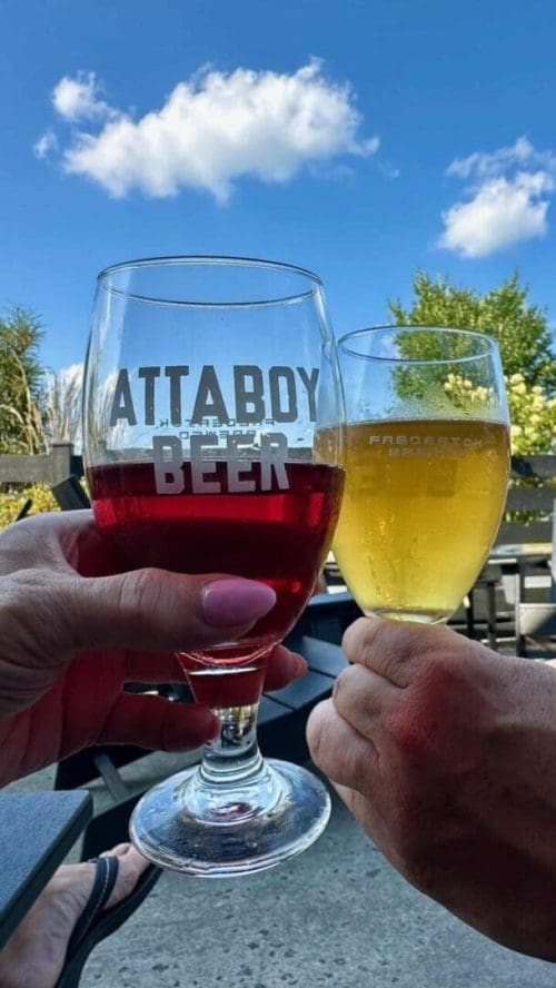 Two people toast with ATTABOY BEER and FARBOOSTER outdoors, enjoying craft brews after exploring the best hikes around Frederick, MD. Blue sky, clouds, trees, and patio furniture create a perfect post-hike backdrop.