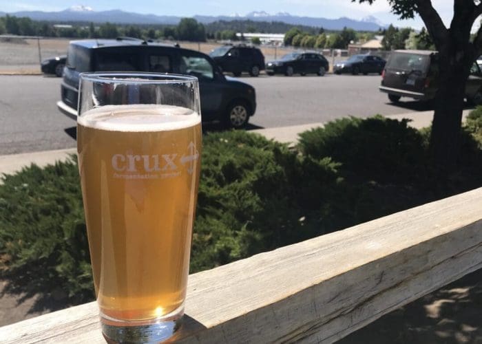 A pint glass of hazy, golden beer from one of the best breweries Bend has to offer sits on a wooden railing outdoors, with greenery, parked cars, and snow-capped mountains beneath a sunny blue sky in the background.