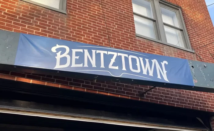 A brick building with two windows displays a blue sign reading BENTZTOWN in white, stylized letters. Below, people are seated at tables inside the warm-lit space—one of the best bars in Frederick—visible through a large open window.