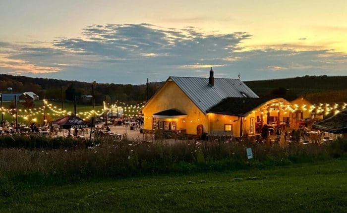 A yellow building with string lights outside glows warmly at sunset. People gather at outdoor tables, enjoying the cozy atmosphere typical of Frederick MD Wineries. Rolling hills and fields surround the scene beneath a cloudy, sunlit sky.
