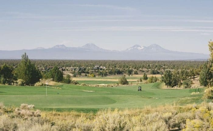 The scenic golf course at Brasada Ranch with lush green fairways and a golf cart sits in the center. Sagebrush and trees surround the course near rolling forests and snow-capped mountains—perfect for those seeking where to stay in Bend, Oregon.