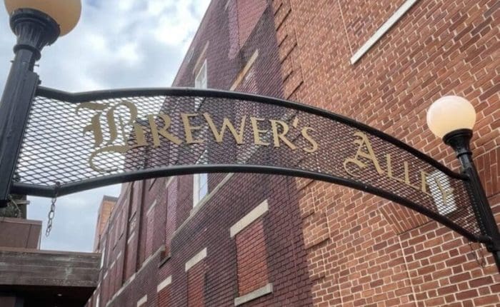 A decorative black iron archway with gold letters reading Brewer’s Alley, one of the best bars in Frederick, stands between two lampposts in front of a tall red brick building. The sky is partly cloudy, and a sidewalk runs alongside the building.