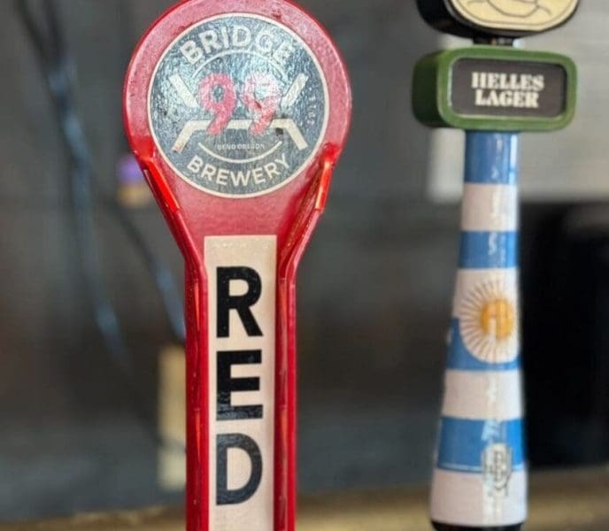 A close-up of two beer tap handles at one of the top breweries in Bend, Oregon. The left handle is red and labeled “RED: BRIDGE BREWERY ROCK CRAWLER.” The right features a cartoon face, green accent, and Argentina flag-inspired stripes.