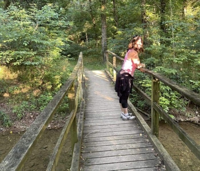 A woman stands on a wooden footbridge in a lush, green forest along one of the best hikes around Frederick MD. She leans on the railing, sunlight filtering through the trees and casting dappled light on the bridge and surrounding foliage.