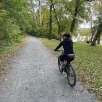 A person wearing a black outfit and cap rides a bicycle on a gravel path through a lush, green forest along one of the scenic bike trails near Frederick MD. Fallen leaves border the path, trees line both sides, and a river is partially visible in the background.