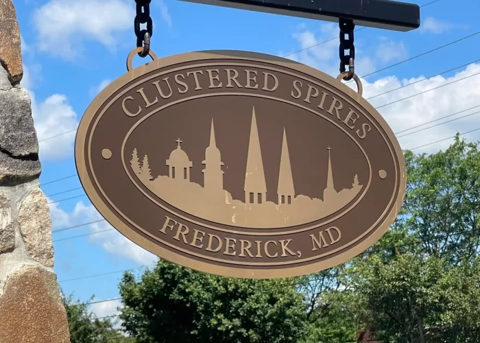 Oval tan sign reading “CLUSTERED SPIRES FREDERICK, MD,” featuring church spires silhouettes, hangs from a metal bracket on a stone pillar. In the background are trees, parked white vans, and a blue sky with clouds near New Market, MD.