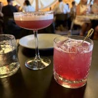 Two pink cocktails sit on a dark table: one is in a coupe glass with foam on top, the other in a short glass with ice and a cocktail pick. A glass of water, an empty plate, and people dining inside a warmly-lit tented space are visible in the background.