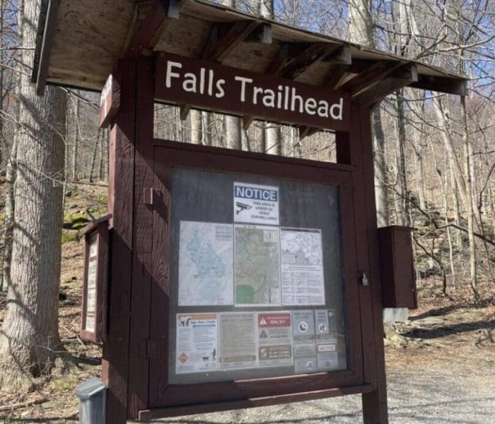 A wooden trailhead sign labeled “Falls Trailhead” stands in a forest with bare trees—one of the best hikes around Frederick MD. The sign displays maps, notices, and safety info inside a glass case as sunlight highlights the gravel-covered ground.