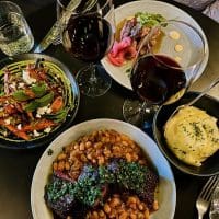 Overhead view of a table set with two glasses of red wine, a plate of stewed beans with braised meat and green sauce, a bowl of creamy mashed potatoes, a dish of roasted carrots topped with mint and feta, and a plate of sliced meat with pickled onions and greens.