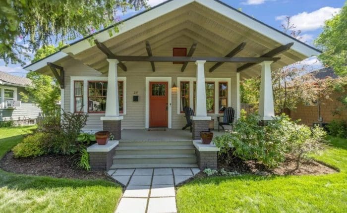 A charming craftsman-style house—an ideal option if you’re wondering where to stay in Bend, Oregon. Enjoy the front porch with white columns, a red door, brown trim, and lush greenery all around under a blue sky dotted with white clouds.