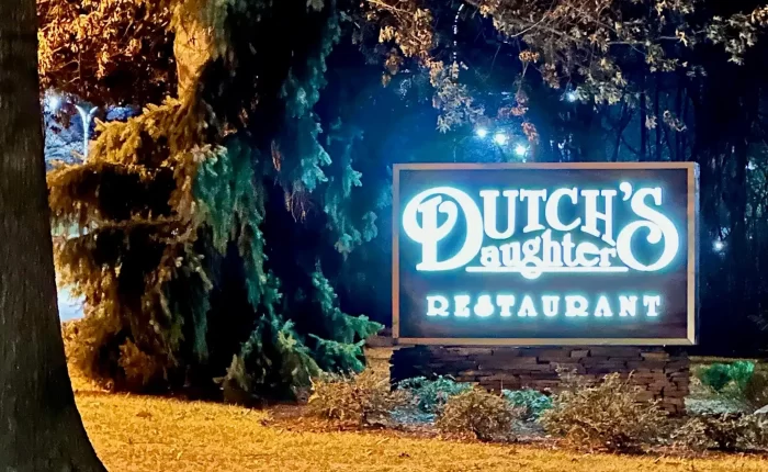 A brightly lit sign at night reads Dutch’s Daughter Restaurant, one of the best bars in Frederick. The sign is surrounded by greenery and trees, with fallen leaves covering the ground and a warm glow shining against the dark, wooded background.