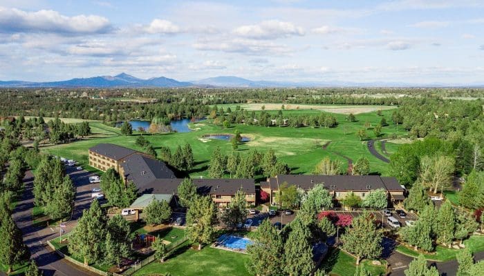 Aerial view of a golf course with green fairways, ponds, and surrounding trees near a hotel building in the foreground.