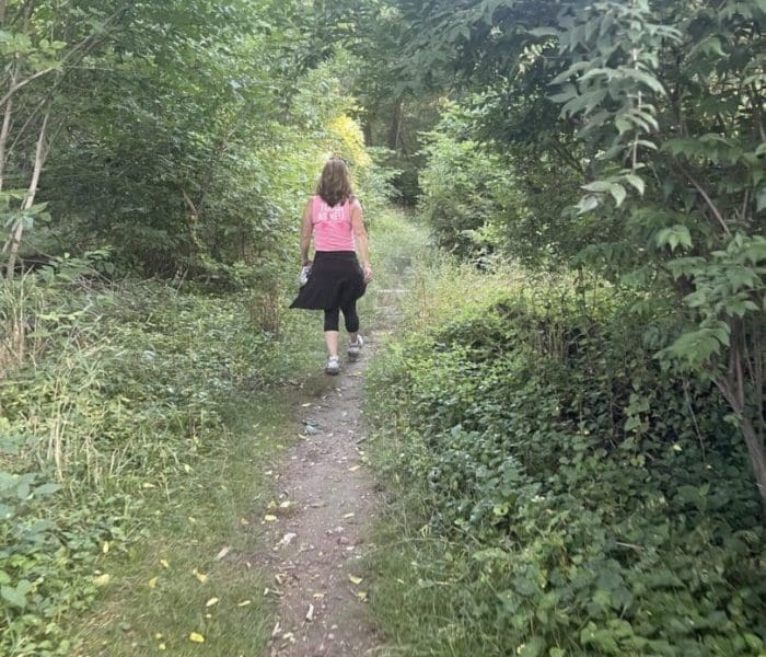 A person with long brown hair, wearing a pink shirt and black pants, walks alone on a narrow dirt path—one of the best hikes around Frederick MD—surrounded by dense green foliage and trees as sunlight filters through the leaves, creating a peaceful scene.