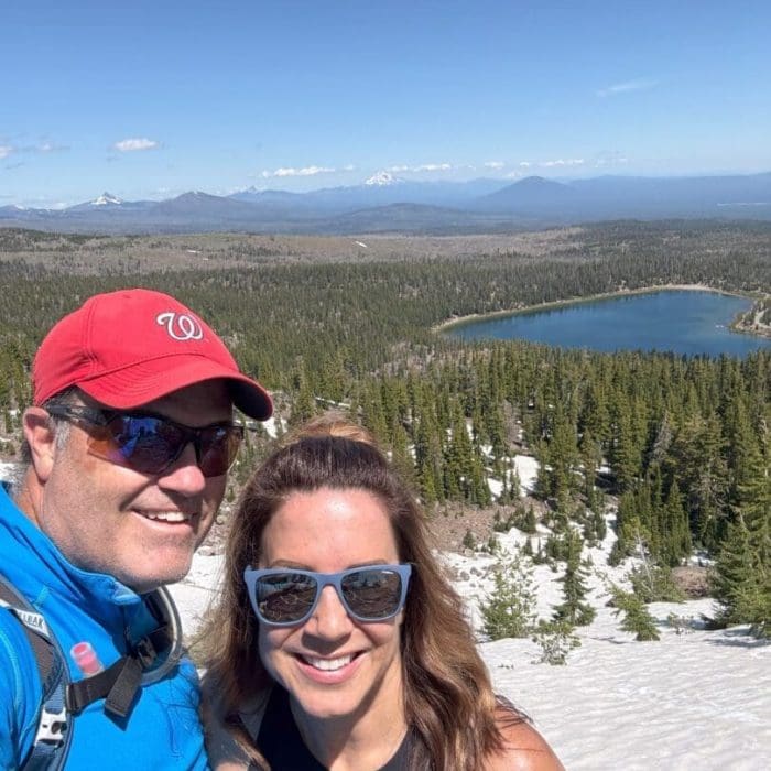 A smiling man and woman wearing sunglasses pose on a sunny, snow-covered mountain during one of their favorite Bend hikes. Behind them are pine trees, a blue lake, and distant mountains under a clear sky.